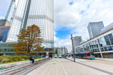 Frankfurt, Germany - October 21, 2025: Modern urban street scene in Frankfurt, Germany, showcasing a towering glass skyscraper, autumn tree, and a moving tram, highlighting dynamic city infrastructure
