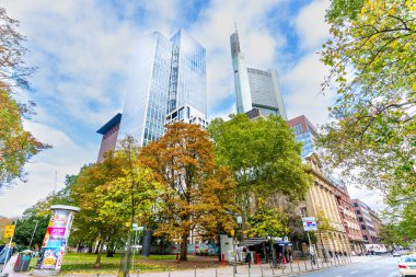 Frankfurt, Almanya - 21 Ekim 2025: Frankfurt 'un modern gökdelenleri parlak mavi gökyüzü altında hareketli bir şehir sokak manzarasını çerçeveleyerek canlı sonbahar yaprakları arasında dururlar.