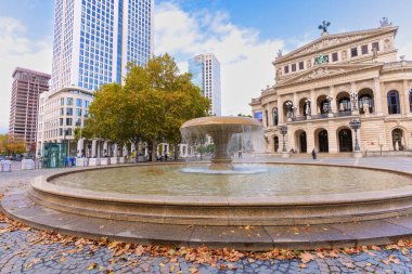 Frankfurt, Germany - October 21, 2025: Historic Alte Oper with a prominent fountain in the foreground, flanked by modern skyscrapers and autumn leaves adorning the cobblestone square on a clear day.
