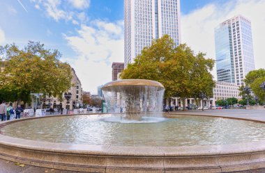 Frankfurt, Germany - October 21, 2025: Frankfurt's vibrant city square showcases a grand fountain and towering skyscrapers amidst autumn foliage under a bright sky.