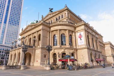 Frankfurt, Germany - October 21, 2025: Wide angle view of the Old Opera House building with ornate details and an active outdoor cafe, juxtaposed with a modern skyscraper under a clear daytime sky.