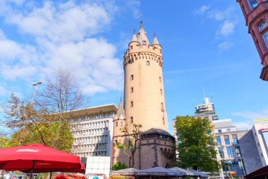 Frankfurt, Almanya - 21 Ekim 2025: Frankfurt 'taki Eschenheimer Kulesi açık mavi bir gökyüzünün altında, aydınlık bir günde modern şehir yapıları ve ağaçlarla çevrili duruyor..