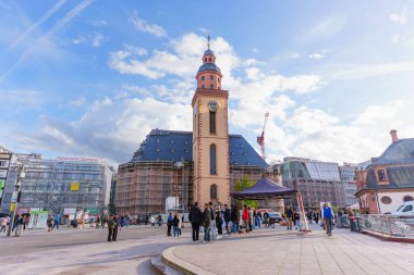 Frankfurt, Germany - October 21, 2025: Frankfurt's historic Katharinenkirche tower stands prominently with scaffolding on a busy Hauptwache square where people gather under a cloudy blue sky.