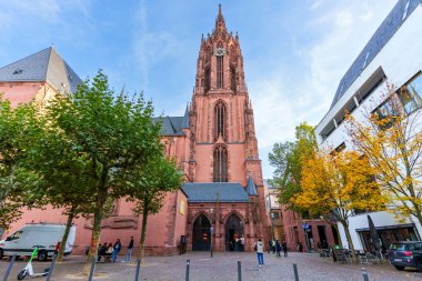 Frankfurt, Almanya - 21 Ekim 2025: Görkemli kırmızı kumtaşı Frankfurt Katedrali, modern binalarla çelişen ve sonbahar yapraklarıyla çerçevelenmiş canlı bir kaldırım taşı plazasını gururla gözden geçirir..