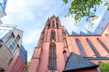 Frankfurt, Almanya - 21 Ekim 2025: Frankfurt Katedrali 'nin görkemli Gotik Kulesi, karmaşık kırmızı kumtaşı detaylarıyla modern binalarla çevrili mavi gökyüzüne karşı görkemli bir şekilde yükseliyor.