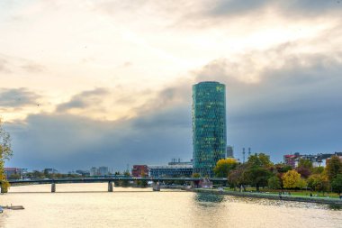 Frankfurt, Almanya - 21 Ekim 2025: Westhafen Kulesi Frankfurt 'un ana nehri üzerinde süzülüyor, modern bir köprü ve dinamik bulutlu bir gökyüzü altında canlı sonbahar ağaçları yer alıyor..