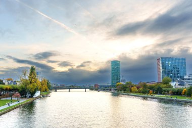 Frankfurt, Almanya - 21 Ekim 2025: Frankfurt 'un canlı Main River sahnesinde modern gökdelenler, bir köprü ve dinamik bir gökyüzü altında popüler gezinti alanı oluşturan sonbahar ağaçları yer almaktadır..