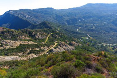 Sarmal toprak yollar geniş Calabasas dağ manzarasını dolaşır, yemyeşil bitki örtüsü, kayalık çıkıntılar ve uzak bir vadide yuva yapmış bir topluluk ortaya çıkar..