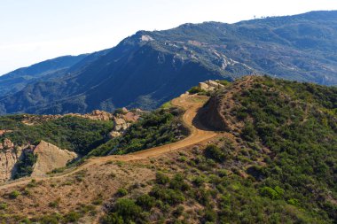 Kurak, engebeli yamaçlar boyunca uzanan sarmal toprak yol yeşil çalılarla kaplı, berrak mavi gökyüzünün altındaki uzak, ormanlık dağlara çıkıyor..