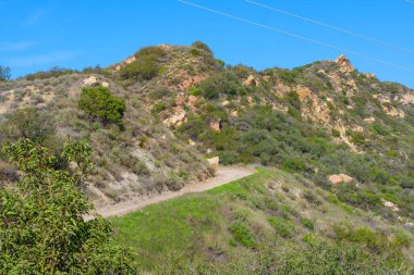 Kıvrımlı toprak yol, şaparal ve kaya oluşumlarıyla örtülü güneşli bir yamaçtan yükselir. Yürüyüşçüleri canlandırıcı bir şehir kaçışı için doğal güzelliği keşfetmeye davet eder..