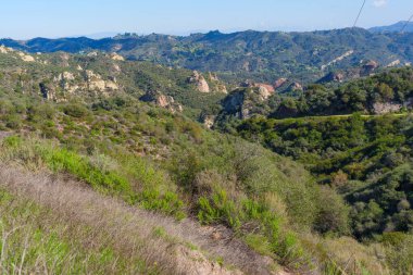 Yayılan manzarada, Kızıl Kaya Kanyon Parkı 'nın engebeli doğal güzelliği eşsiz kaya oluşumları, yeşil şaparral ve açık gökyüzünün altındaki uzak mavi tepeler sergilenmektedir..