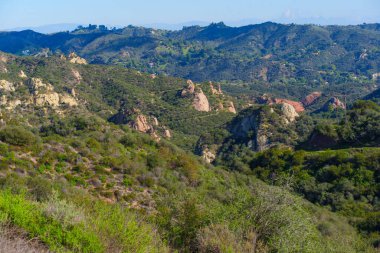 Panoramik manzara, açık mavi gökyüzünün altında eşsiz kırmızı ve altın kaya oluşumlarına sahip yemyeşil yamaçları gösteriyor. Kızıl Kaya Kanyon Parkı 'nın yürüyüş ve aktif yaşam tarzı için engebeli güzelliğini vurguluyor..
