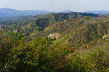 Genişletici görüntü dalgalı yeşil tepeler ve Red Rock Canyon Parkı 'ndaki kayalık araziyi ortaya çıkarıyor. Uzak yapılar, elektrik hatları ve açık bir gökyüzü sergiliyor..