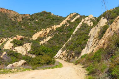 Dolambaçlı toprak yol, güneşten sırılsıklam olmuş yamaçlardan geçiyor parlak mavi gökyüzünün altındaki yeşil şapel arasında altın kaya oluşumları sergiliyor..