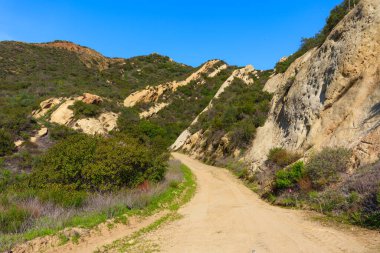 Kırmızı Kaya Kanyon Parkı 'ndan yükselen sarmal toprak yol, güneşli, engebeli kaya oluşumları ve parlak mavi gökyüzü altında canlı yeşil şapellerle çevrili..