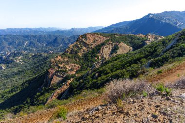 Yüksek görüş açısına sahip engebeli Kaliforniya manzarası dramatik kırmızı kaya oluşumlarını, yemyeşil şapeli ve berrak gökyüzünün altındaki uzak dağları gözler önüne seriyor..