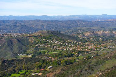 Çok sayıda evi ve yeşil ağaçları olan geniş yerleşim yerleri berrak mavi bir gökyüzünün altında, yuvarlanan, fırçayla kaplı tepelerden oluşan bir vadi boyunca esiyor..