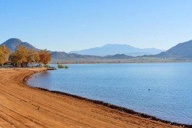 Perris Gölü 'nün pırıl pırıl mavi suları kıyı şeridiyle buluşuyor, sonbahar ağaçları ve açık mavi gökyüzünün altındaki uzak dağlar..