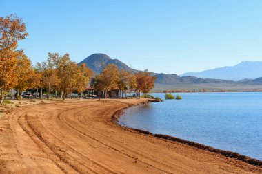 Perris Gölü manzarası, sakin suların yanında, altın sonbahar ağaçları ve engebeli dağ zirveleriyle çevrelenmiş eğri bir toprak yol sergiliyor..