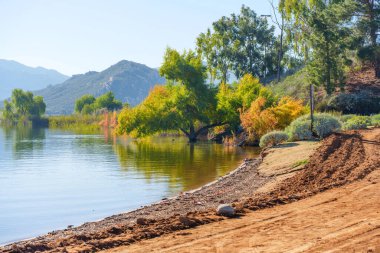 Perris Gölü 'nün sakin suları parlak, berrak bir gökyüzünün altındaki yuvarlanan tepelere doğru uzanan verimli bir sonbahar yeşilliğini ve iyi yıpranmış bir toprak yolu yansıtır..