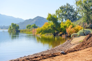 Perris Gölü 'nün sakin suları güneşli kıyılarındaki canlı ağaçları yansıtır. Uzak dağlar berrak gökyüzünün altında dingin bir zemin oluşturur..
