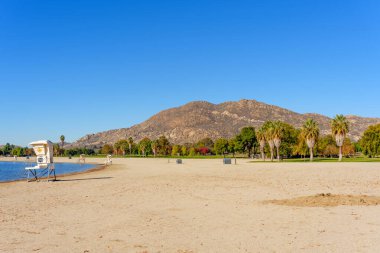 Perris Gölü 'nü çevreleyen geniş kumlu sahilde beyaz cankurtaran duruyor. Kayalık dağlar ve parlak mavi gökyüzünün altında görünen çeşitli ağaçlar..