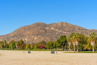 Perris Gölü 'nün parlak mavi gökyüzünün altında geniş kumlu bir ön planda, engebeli engebeli dağlık zemin çeşitli yeşil ve sonbahar ağaçlarını büyütür..