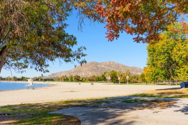 Berrak mavi gökyüzünün altında Perris Gölü 'nün kumlu plajında sonbahar renklerinde ağaçlar ve engebeli dağ zeminleri var..