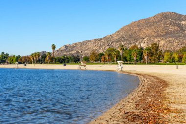 Perris Gölü 'nün mavi suları, berrak bir gökyüzünün altında engebeli bir dağ tarafından desteklenen cankurtaran kuleleri ve ağaçlarla dolu kumlu bir sahille buluşur..