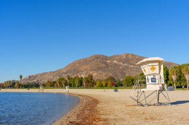 Perris Gölü 'nün manzaralı sahili beyaz cankurtaran kuleleri, durgun mavi su, yeşil ve sonbahar ağaçlarının bir karışımı ve berrak mavi gökyüzünün altında engebeli dağlar içerir..