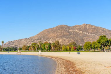 Perris Gölü 'nün kumlu kıyı şeridi, berrak bir gökyüzünün altında, sonbahar ağaçları ve engebeli bir dağ tarafından çerçevelenmiş sakin mavi suyun yanında uzanır..