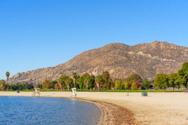 Perris Gölü 'nde güneşli bir gün. Huzurlu suları, kumlu plajı, iki cankurtaran standı, canlı ağaçlar ve yüksek bir kayalık dağ..