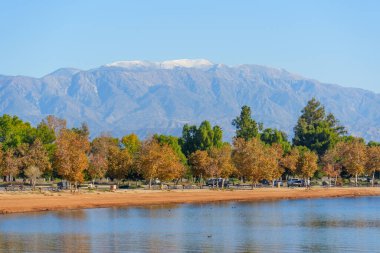 Titrek sonbahar ağaçları, Perris Gölü 'nün kumlu kıyısı hizasında; sakin suları, uzaktaki görkemli karla kaplı dağlarıyla berrak mavi bir gökyüzünü yansıtır..
