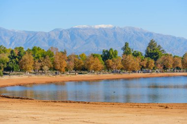 Parlak sonbahar ağaçları Perris Gölü 'nün kumlu kıyılarında uzanır. Arka planda açık mavi bir gökyüzünün altında yükselen görkemli karlı dağlar vardır..