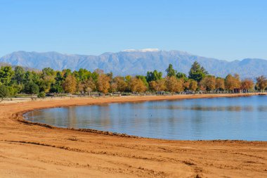 Perris Gölü Eyalet Dinlenme Alanı, berrak bir gökyüzü altında sakin mavi sular, geniş bir kumlu bir sahil, sonbahar yaprakları ve karla kaplı dağlar sergiliyor..