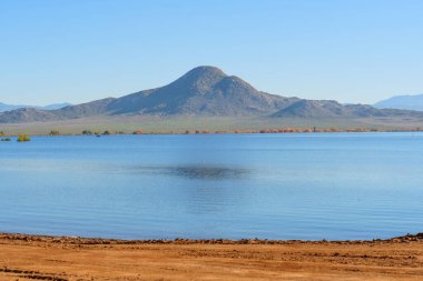 Geniş Perris Gölü suları berrak mavi bir gökyüzü altında görkemli engebeli dağları yansıtıyor, kırsal bir kıyı şeridi ve uzak sonbahar yapraklarıyla.