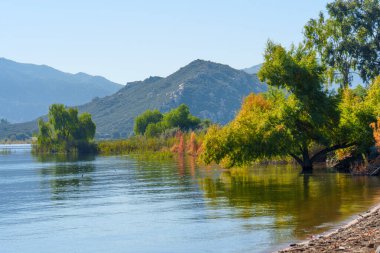 Perris Gölü 'nün sakin mavi suları, uzaktaki engebeli dağlarla çevrili yeşil ve altın ağaçlarla çevrili sakin gökyüzünü yansıtır..