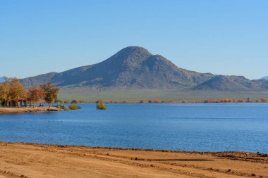 Perris Gölü 'nün geniş manzarası engebeli dağlar, durgun mavi su boyunca sonbahar renkli ağaçlar ve berrak, parlak bir gökyüzü içerir..