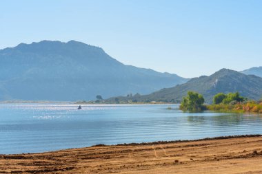 Işıldayan Perris Gölü suları berrak mavi bir gökyüzünü yansıtır, uzak puslu dağlar ve tek bir su aracı, ham, dokulu bir toprak ön planı ile.