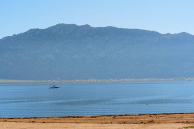 Perris Gölü 'nün engin genişliği berrak bir mavi gökyüzü altında parıldıyor. Uzaktan sisli dağlarla çevrili, tek başına bir tekneye ev sahipliği yapıyor..