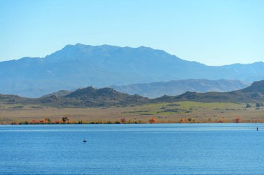 Perris Gölü, sonbahar renklerinde kıyı şeridi, engebeli tepeler ve uzak puslu dağlarla çevrili berrak bir gökyüzünü yansıtan sakin mavi suları sunar..