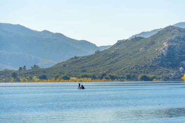Küçük bir teknede iki kişi Perris Gölü 'nün durgun mavi sularında dolaşır. Berrak bir gökyüzünün altındaki yemyeşil dağlarla çevrilidir..