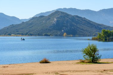 Berrak bir gökyüzünün altında engebeli, yeşil kaplı dağlarla desteklenmiş iki kişilik küçük bir balıkçı teknesinin yer aldığı sakin bir mavi su alanı..