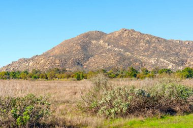 Engebeli dağ, berrak mavi gökyüzü altında engin doğal manzaraya hükmediyor. Önplanda kuru altın otlar ve dağınık yeşillik var..