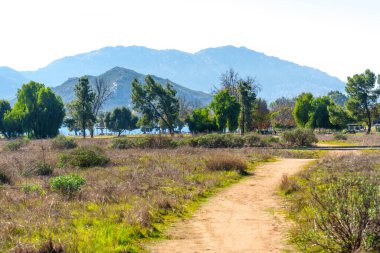 Tozlu patika, ağaçlarla ve uzak puslu dağlarla çevrili mavi bir göle uzanan yeşil ve kahverengi bitkilerle kurak bir arazide yol alır..