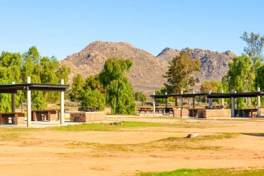 Perris Gölü 'ndeki piknik sahalarında çeşitli gölgeli masalar, yeşil ağaçlar ve parlak, berrak bir gökyüzünün altında dağlar var..