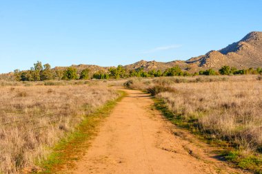 Kuru altın çim tarlalarından yeşil ağaçlara ve engebeli dağlara uzanan sarmal toprak patikalar Perris Gölü dinlenme bölgesinde berrak mavi bir gökyüzü altında.