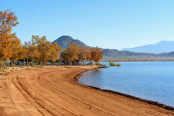 Perris Gölü manzarası, sakin suların yanında, altın sonbahar ağaçları ve engebeli dağ zirveleriyle çevrelenmiş eğri bir toprak yol sergiliyor..
