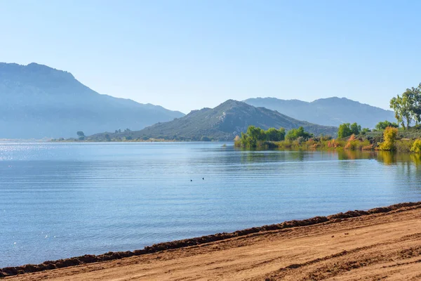 Perris Gölü 'nün sakin mavi suları berrak bir gökyüzü altında parıldıyor, sisli uzak dağlar ve canlı bir sonbahar kıyı şeridi tarafından kuşatılmış..