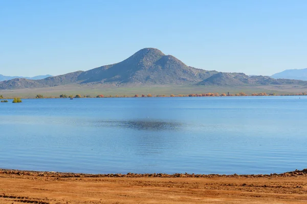 Geniş Perris Gölü suları berrak mavi bir gökyüzü altında görkemli engebeli dağları yansıtıyor, kırsal bir kıyı şeridi ve uzak sonbahar yapraklarıyla.